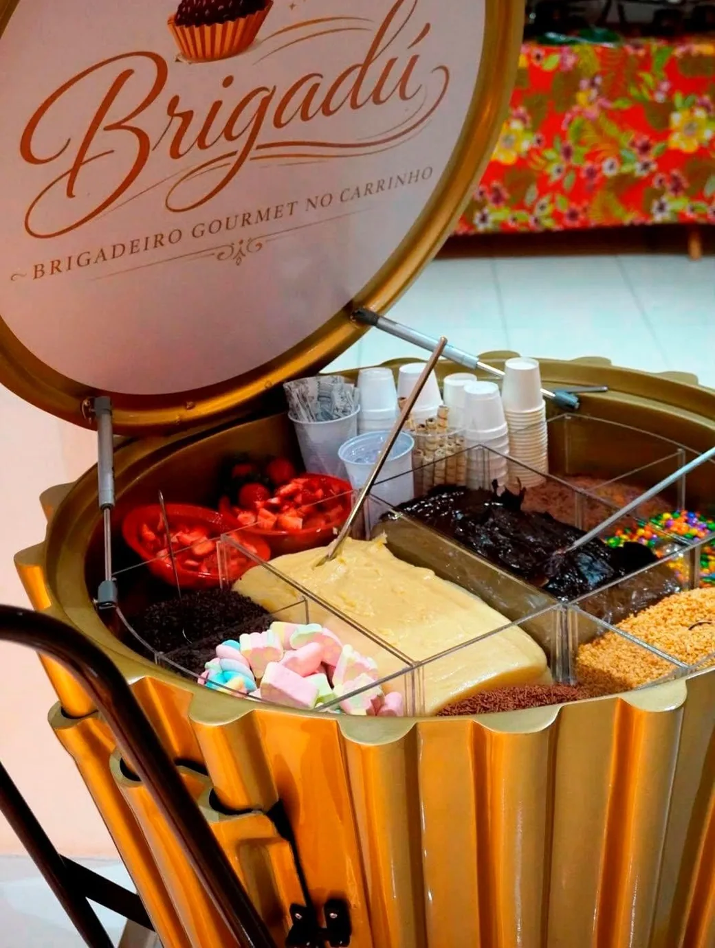 Detalhe do corpo dourado canelado do carrinho Brigadú aberto, com as cubas de brigadeiro e toppings à mostra